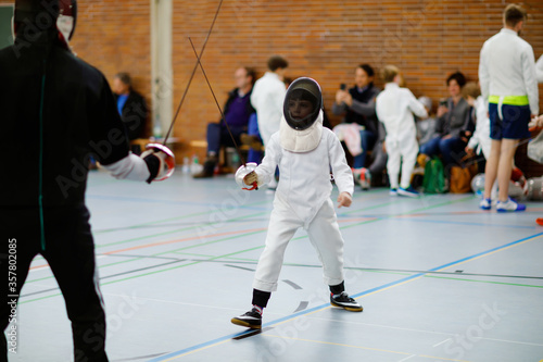 Little kid boy fencing on a fence competition. Child in white fencer uniform with mask and sabre. Active kid training with teacher and children. Healthy sports and leisure. © Irina Schmidt