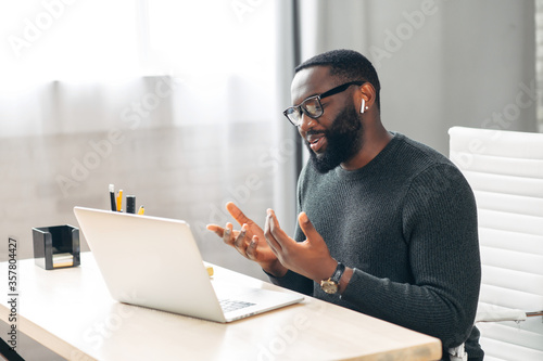Side view of African-American office employee talking via video connection while sitting at the desk. A guy using laptop for video call in contemporary office space