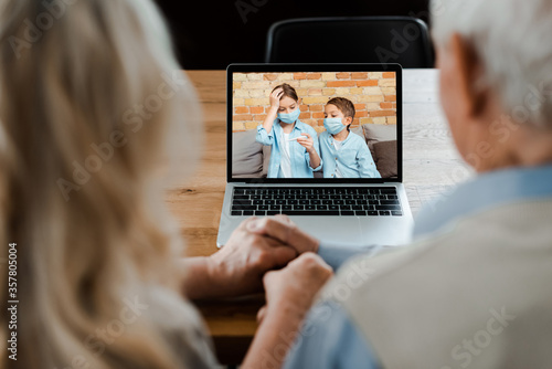 grandparents holding hands and having video chat with grandchildren in medical masks holding thermometer during self isolation