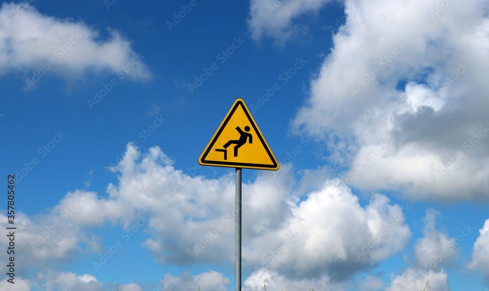 Yellow triangular falling danger sign against blue sky with fluffy clouds on background Yellow triangular falling danger sign against blue sky with fluffy clouds on background