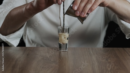 The bartender pours irish cream from jigger to cocktail glass with sambuca