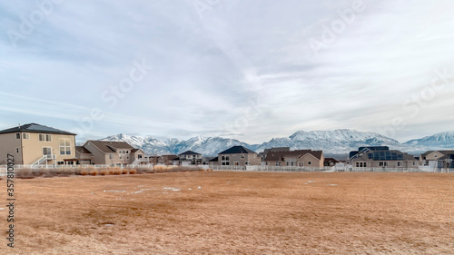 Panorama crop Grassy field with melting snow against homes and snowy mountain background