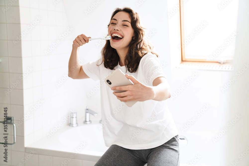 Photo of cheerful woman cleaning teethes while using mobile phone Photo of cheerful woman cleaning teethes while using mobile phone