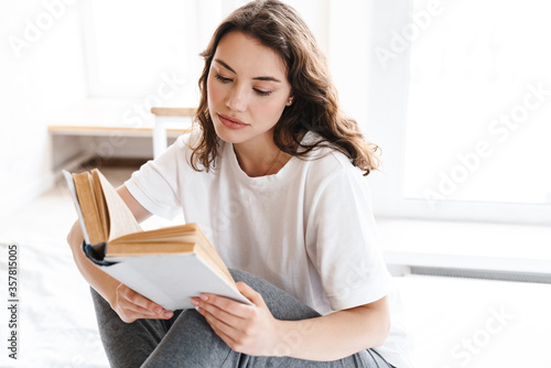 Photo of focused attractive woman reading book while sitting on bed