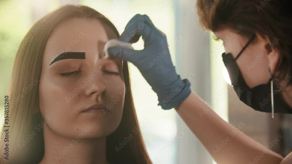 Cosmetologist wipes out the tinted color from the brow of a young woman Cosmetologist wipes out the tinted color from the brow of a young woman