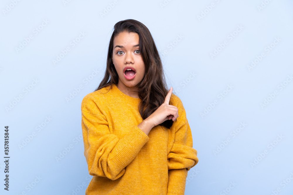 Young Colombian girl with sweater over isolated blue background surprised and pointing side Young Colombian girl with sweater over isolated blue background surprised and pointing side