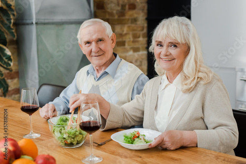 smiling senior couple having dinner with wine and salad at home on self isolation