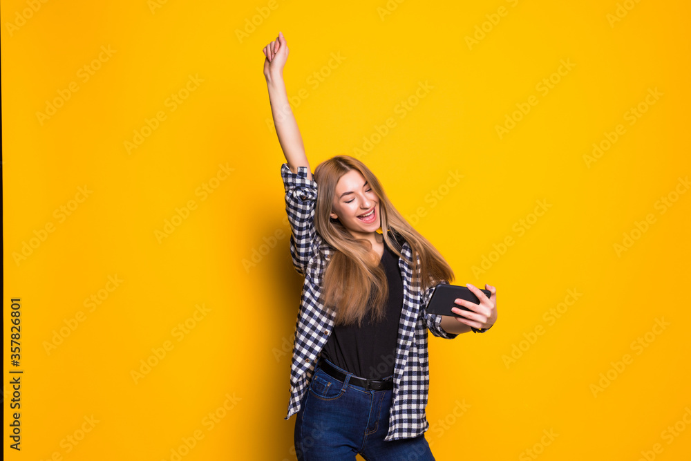Portrait of a cheerful young woman holding mobile phone, celebrating standing isolated over yellow background