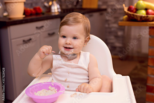 A happy baby 10-12 months old eats milk porridge with a spoon. Portrait of a happy girl in a highchair in the kitchen