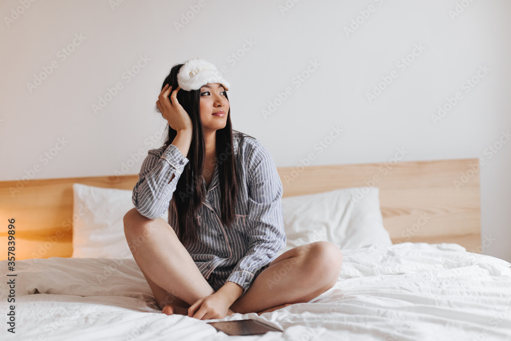 Girl with dark long hair is resting on white bed. Portrait of woman in pajamas and sleep mask Girl with dark long hair is resting on white bed. Portrait of woman in pajamas and sleep mask