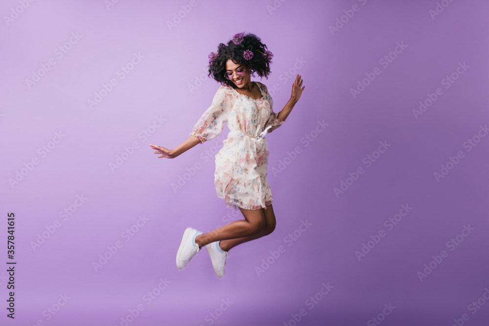 Carefree african girl in white shoes jumping in studio. Adorable female model with flowers in hair dancing on purple background with happy smile.