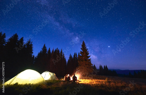 Night camping with bonfire. People bask in campfire near illuminated tent city, enjoying valley of mountains in pine forest. Dark blue night sky is strewn with bright stars and Milky Way