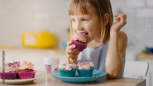 In the Kitchen  Adorable Little Girl Eats Creamy Cupcake with Frosting and Sprinkled Funfetti. Cute Hungry Sweet Tooth Child Bites into Muffin with Sugary Frosting
