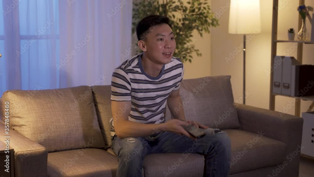 chinese young man slacking off after work watching amusing tv program in living room. asian male holding remote control sitting at home is interested in the content of show and turning up volume. chinese young man slacking off after work watching amusing tv program in living room. asian male holding remote control sitting at home is interested in the content of show and turning up volume.