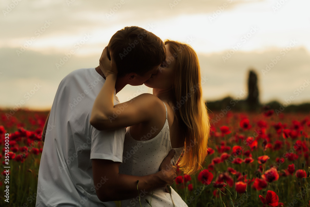 Sensual portrait of a young couple kissing in poppy field. Romantic couple in a poppy field standing in an embrace on a summer day at sunset. Copy space.