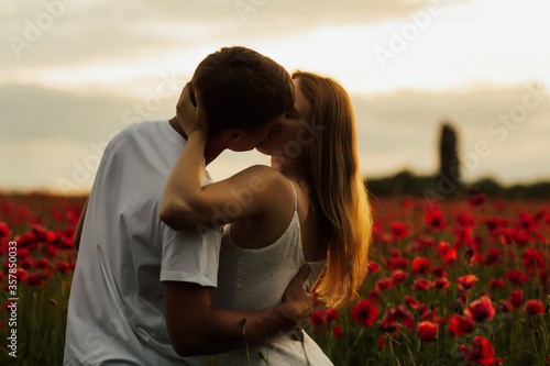 Sensual portrait of a young couple kissing in poppy field. Romantic couple in a poppy field standing in an embrace on a summer day at sunset. Copy space.