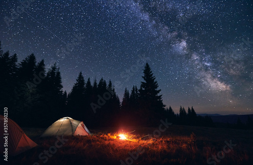 Burning bonfire in tent city without people under bright starry sky with Milky way against the backdrop of silhouettes pine forest and valley of hills. Healthy lifestyle and relaxation concept.