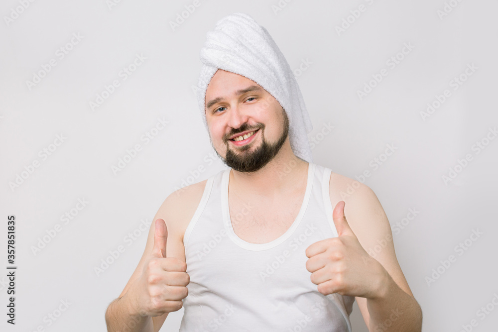 Happy smiling Caucasian bearded man in whilte t-shirt and with a towel on his head posing to camera on isolated while background and showing his thumbs up. Spa procedures for men Happy smiling Caucasian bearded man in whilte t-shirt and with a towel on his head posing to camera on isolated while background and showing his thumbs up. Spa procedures for men