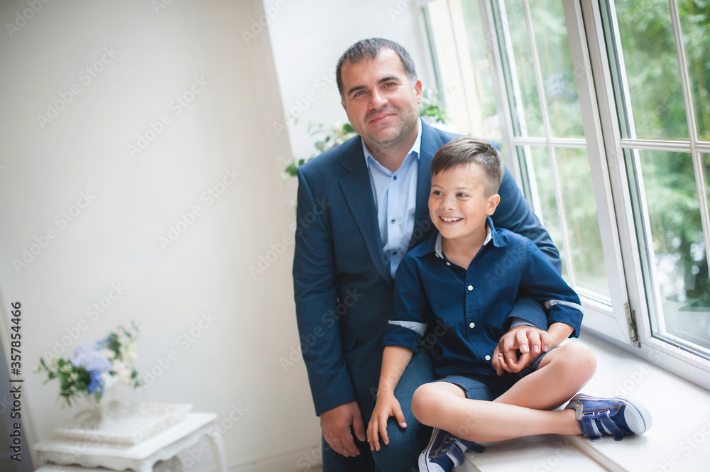 A man in a blue suit sits on a windowsill with his son. Solemn event, smiling. Beautiful and cheerful. A man in a blue suit sits on a windowsill with his son. Solemn event, smiling. Beautiful and cheerful.