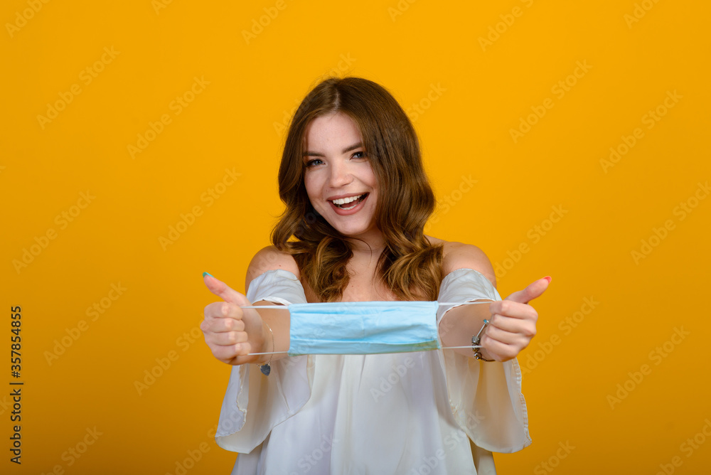 Beautiful happy young woman with face mask in hands on yellow background. COVID virus and air pollution. Recovering from illness, end of epidemic. Beautiful happy young woman with face mask in hands on yellow background. COVID virus and air pollution. Recovering from illness, end of epidemic.