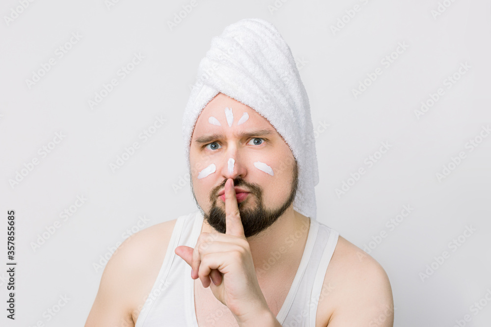 Funny crazy bearded man with a white towel on his head and cream on face, looking at camera on white background and saying hush be quiet with finger on lips gesture Funny crazy bearded man with a white towel on his head and cream on face, looking at camera on white background and saying hush be quiet with finger on lips gesture