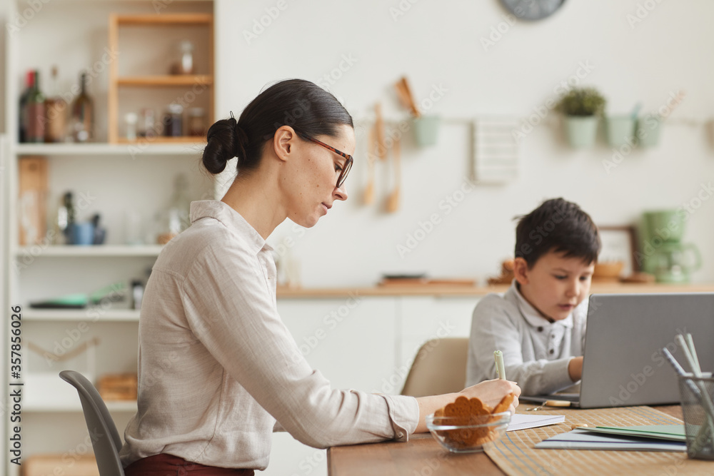 Side view portrait of young mother helping son studying at home while sitting at desk and writing in cozy interior, copy space Side view portrait of young mother helping son studying at home while sitting at desk and writing in cozy interior, copy space
