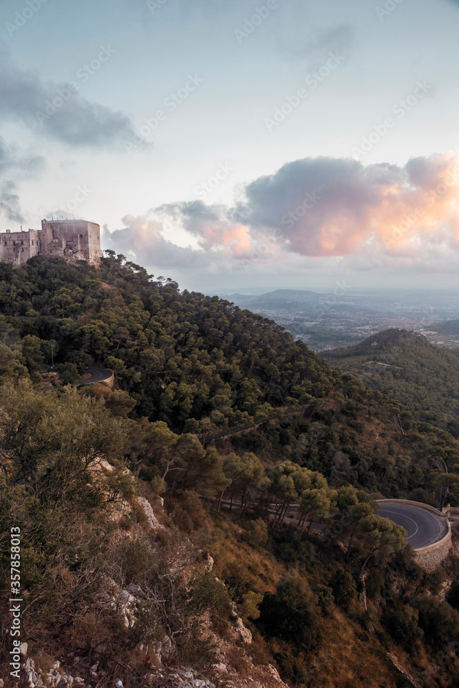 Obraz premium summer afternoon shot of the Puig de Maria sanctuary and monastery standing at the top of the green Sant Marti hills with beautiful mountains and a blue sky with white pink clouds in the back