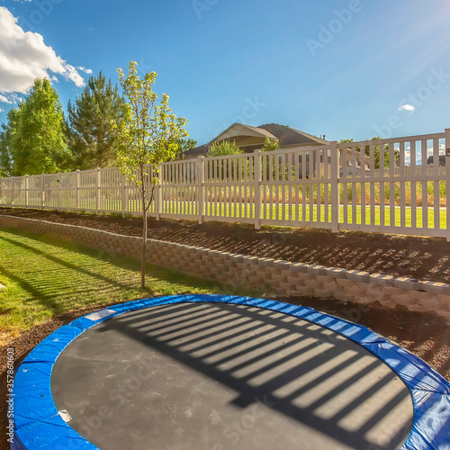 Square crop Trampoline at sunlit backyard of home with patio and planting bed against fence