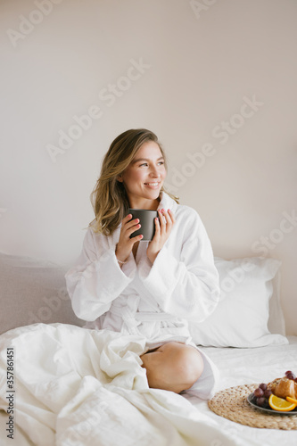 Фотография Young beautiful woman wearing white bathrobe having breakfast in bed with coffee and croissant and fresh fruits in cozy bedroom