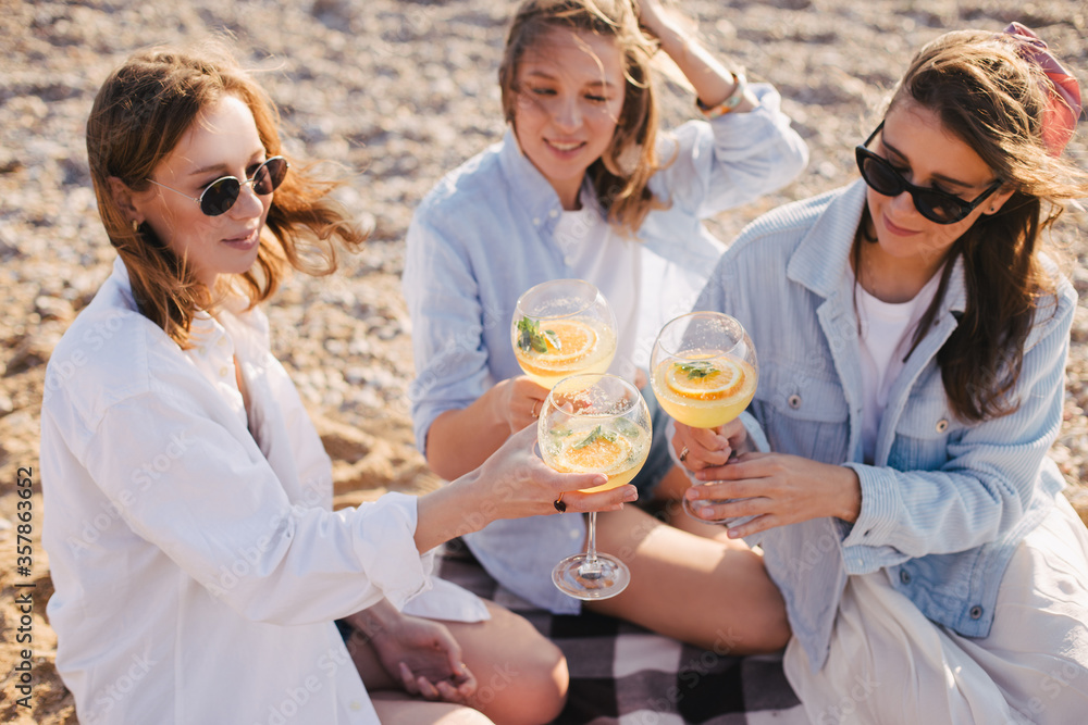 Three young beautiful happy women female friends having cozy summer picnic with lemonade, fresh bread and fruits on a beach. Three young beautiful happy women female friends having cozy summer picnic with lemonade, fresh bread and fruits on a beach.