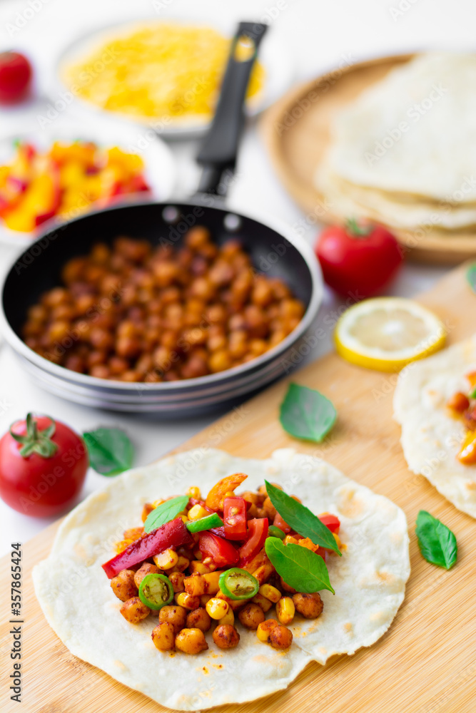 Vegan tortilla tacos with chickpeas, bell peppers, corn, tomatoes, basil leaves and green chilli. On the background are ingredients, lemons, chickpeas in a small pan