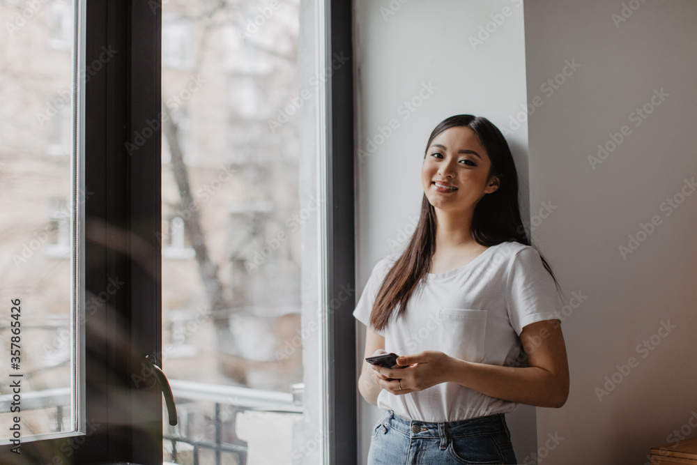 Young woman in white T-shirt leans on wall by window. Girl holds phone and looks into camera with smile