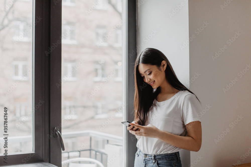 Adorable Asian girl dressed in light top with smile holds smartphone and poses near window Adorable Asian girl dressed in light top with smile holds smartphone and poses near window