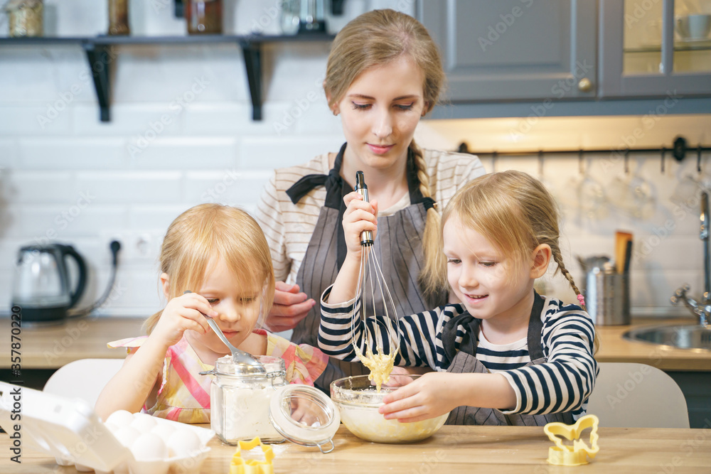 Happy family young mother and two little daughters making dough for pancakes or cookies together in the kitchen Happy family young mother and two little daughters making dough for pancakes or cookies together in the kitchen