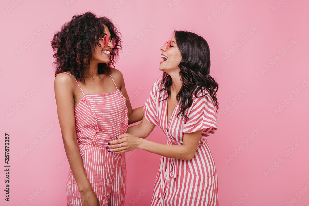 Girl with amazing smile tells her friend funny story. Indoors portrait of models with tanned skin on pink background Girl with amazing smile tells her friend funny story. Indoors portrait of models with tanned skin on pink background