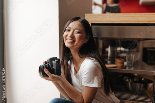 Smiling girl in white t-shirt posing with camera in her hands in kitchen
