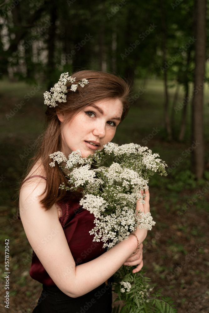 Naklejka premium beautiful red-haired girl hugs a bouquet of wildflowers, stands among the forest thicket, large portrait with freckles