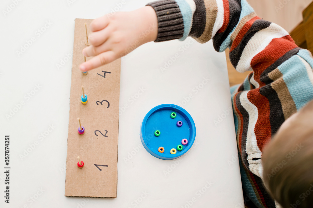 Home made montessori metodic tool for early education, learning to count. 2 year old boy puts beads on wooden rods. Children, people, infancy and education concept. Formation and development of the ch Home made montessori metodic tool for early education, learning to count. 2 year old boy puts beads on wooden rods. Children, people, infancy and education concept. Formation and development of the ch