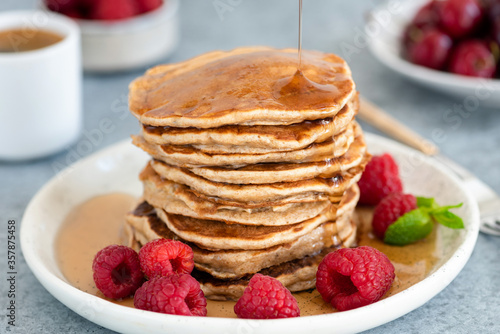 Syrup pouring on stack of whole wheat pancakes served with fresh raspberries on a plate. Closeup view. Healthy breakfast food