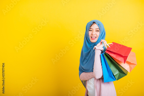 Asian Muslim Arab, Portrait of happy beautiful young woman Islam religious wear veil hijab funny smile she holding colorful shopping bags so glad shopping bags hand raise in studio isolated on yellow