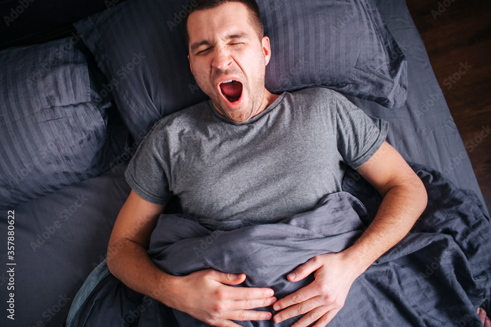 Fototapeta premium Sleepy man in the bed yawns. View from above. Male model wakes up early in the morning.