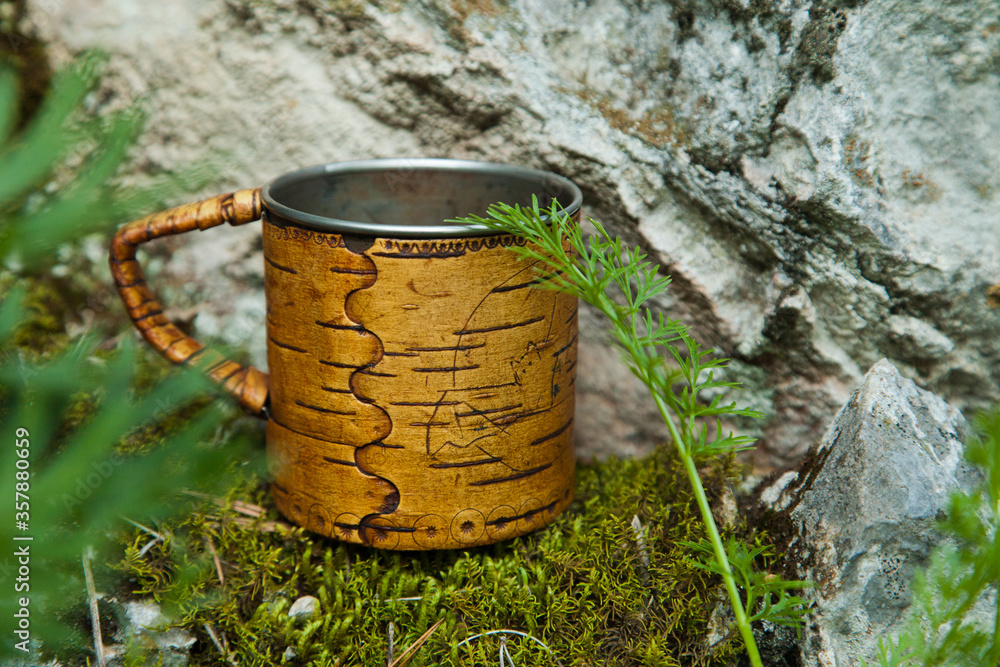 A steel mug with birch bark trim stands on the rocks.