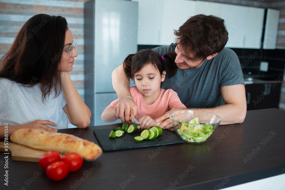 Happy family is cooking in the kitchen. Father teaches daughter to cut vegetables. Mother looks at them Happy family is cooking in the kitchen. Father teaches daughter to cut vegetables. Mother looks at them