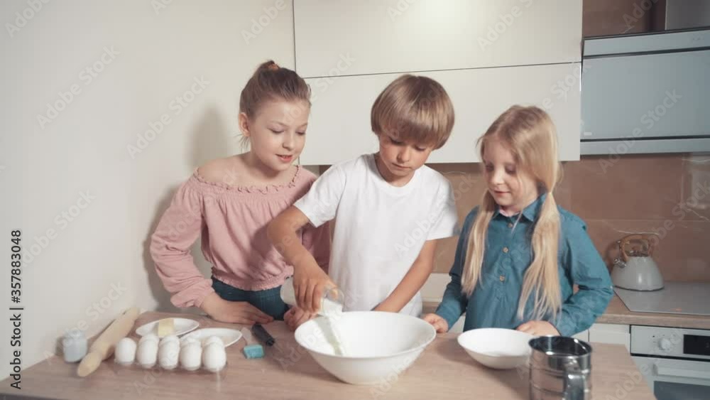 Three children help with cooking in the kitchen. A boy pours milk into a plate. Three children help with cooking in the kitchen. A boy pours milk into a plate.
