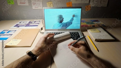 POV shaky camera shot of male student watching online computer programming class with senior teacher explaining how to code on laptop screen. Student taking notes in notepad sitting at desk at home