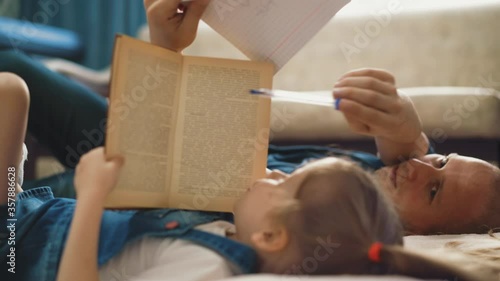 CU, Side view, concept of distance learning: little girl and her bald unshaven dad lie on floor, father knocks on book, which kid covered face, woke up, and shows in notebook on task, closeup photo