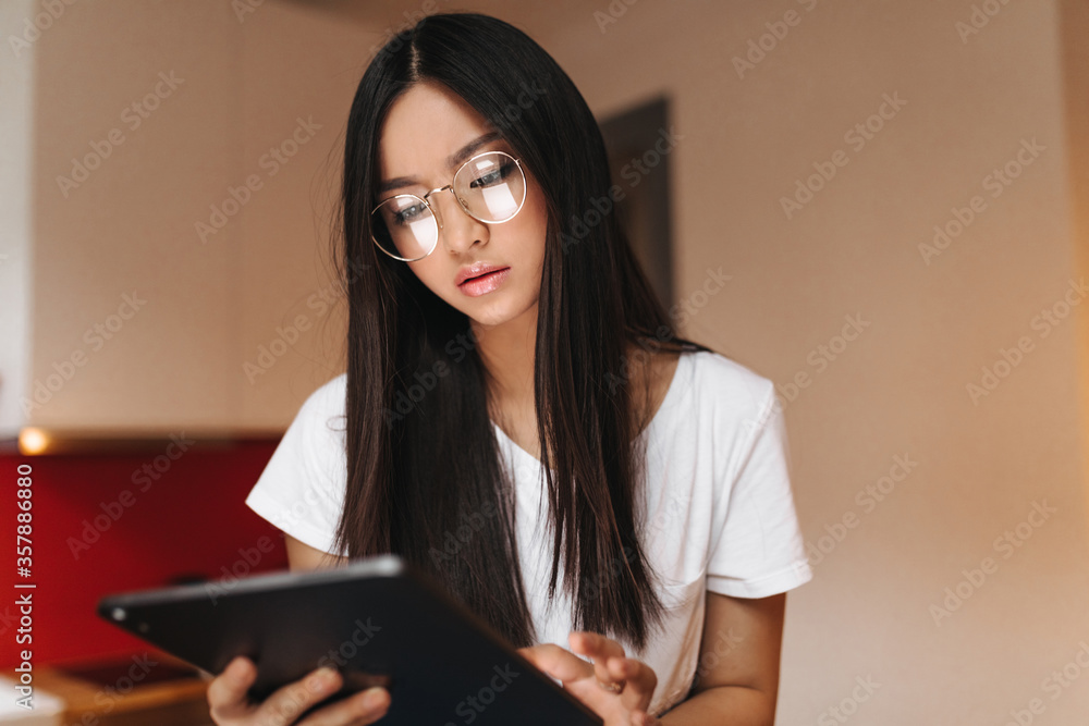 Pensive Asian girl looks into screen of tablet. Brunette woman in glasses posing in kitchen Pensive Asian girl looks into screen of tablet. Brunette woman in glasses posing in kitchen