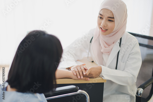 Young Asian woman Muslim doctor giving advice discussion and check up to elderly patient sitting in Wheelchair at hospital which smiling and felling happy. Medicine and health care safe concept.