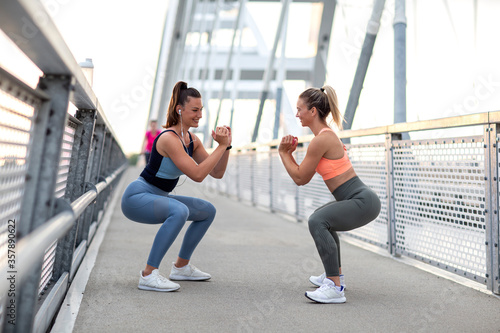 Two girl exercise with weights on hands at bridge at the end of day on sunset