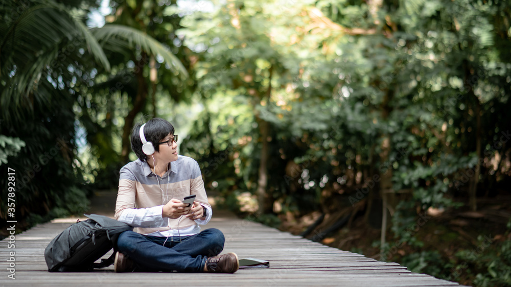 Young Asian man with headphones, glasses and backpack sitting on wooden bridge relaxing in the park. Male university student listening to music using smartphone in the garden. Young Asian man with headphones, glasses and backpack sitting on wooden bridge relaxing in the park. Male university student listening to music using smartphone in the garden.